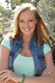 A young blonde woman outside wearing a sleeveless denim jacket and a light-blue top