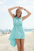 A young blonde woman on a beach wearing a light-blue summer dress