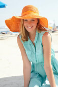 Junge blonde Frau im hellblauen Sommerkleid und orangenem Sommerhut am Strand