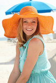 Junge blonde Frau im hellblauen Sommerkleid und orangenem Sommerhut am Strand