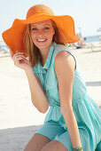 Junge blonde Frau im hellblauen Sommerkleid und orangenem Sommerhut am Strand