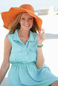 A young blonde woman on a beach wearing a light-blue summer dress and an orange summer hat