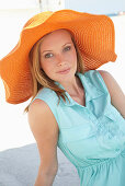 A young blonde woman on a beach wearing a light-blue summer dress and an orange summer hat