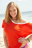 A young blonde woman on a beach wearing a purple bikini and an orange top