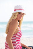 A young blonde woman on a beach wearing a red-and-white striped top and a white hat