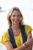 A young blonde woman on a beach wearing a yellow top and a denim waistcoat