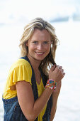 A young blonde woman on a beach wearing a yellow top and a denim waistcoat