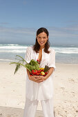 A young brunette woman on a beach wearing a white summer dress and holding a bowl of vegetables