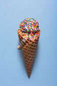 A melting ice cream with colourful sugar sprinkles in a waffle cone, in front of a blue background