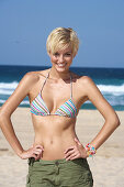 A blonde woman with short hair on a beach wearing a bikini top and shorts