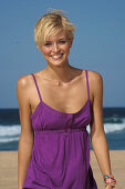 A blonde woman with short hair on a beach wearing a purple top