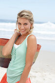 A mature blonde woman on a beach wearing a white top and a turquoise vest with a wicker basket