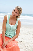 A mature blonde woman on a beach wearing a white top, a turquoise vest and a salmon coloured skirt