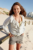 A brunette woman on a sandy beach wearing a light blouse and shorts