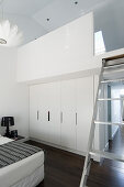 Weißer Einbauschrank, darüber Galerie mit Leiter in weißem Schlafzimmer