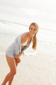 Blonde Frau in weißem Bikini und heller Strickjacke am Strand