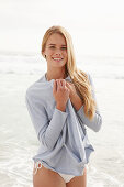 A blonde woman on a beach wearing a white bikini and a light cardigan