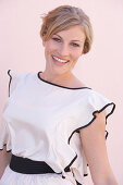 A blonde woman standing against a pink background wearing a light blouse with a black trim