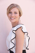 A blonde woman standing against a pink background wearing a light blouse with a black trim