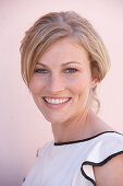 A blonde woman standing against a pink background wearing a light blouse with a black trim