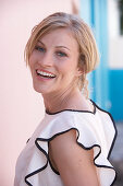 A blonde woman standing against a pink background wearing a light blouse with a black trim