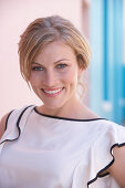 A blonde woman standing against a pink background wearing a light blouse with a black trim