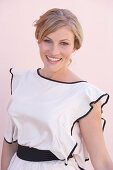 A blonde woman standing against a pink background wearing a skirt and a light blouse with a black trim