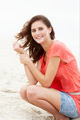 A brunette woman on a beach wearing a salmon-coloured top and a denim skirt