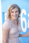 A blonde woman standing against a blue surface wearing a pink short-sleeved blouse