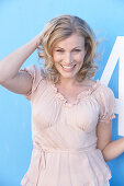 A blonde woman standing against a blue surface wearing a pink short-sleeved blouse