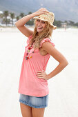 A young blonde woman on a beach wearing a pink top, a short denim skirt and a beige hat