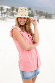 Junge blonde Frau im rosa Top,  kurzem Jeansrock und beigem Hut am Strand