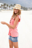 Junge blonde Frau im rosa Top, kurzem Jeansrock und beigem Hut am Strand