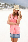 A young blonde woman on a beach wearing a pink top, a short denim skirt and a beige hat