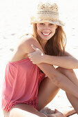 A young blonde woman on a beach wearing a pink top and a beige hat