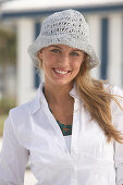 A young blonde woman on a beach wearing a white blouse and a white crocheted hat