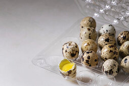 Raw uncooked quail eggs whole and broken in plastic boxing on white marble kitchen table
