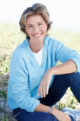 A mature woman with short blonde hair in the countryside wearing blue jeans and a blue jumper over a white shirt
