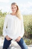 A young brunette woman kneeling in the sand wearing a white blouse and jeans