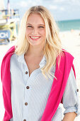 A young blonde woman on a beach wearing a striped shirt with a pink jumper over her shoulders