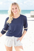 A young blonde woman on a beach wearing a long-sleeved blue shirt and shorts