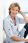 A mature woman with short blonde hair on a beach wearing a light-blue striped short and blue jeans