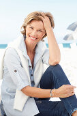 A mature woman with short blonde hair on a beach wearing a light-blue striped short, jumper over her shoulders and blue jeans