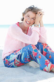 A mature woman with short blonde hair on a beach wearing a pink and white top and colourful trousers