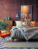 Young woman on double bed with colorful bedding in front of wooden wall