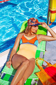 A young blonde woman wearing an orange bathing suit with a bikini top and a hat sitting on a pool lounger