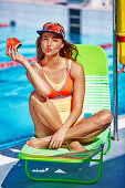 A young blonde woman sitting on a lounger by a pool with a slice of watermelon