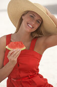 Junge blonde Frau im roten Sommerkleid und mit Sommerhut hält ein Stück Wassermelone