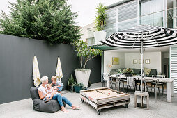 Mother and child sitting on beanbag, sandpit made from old pallets, dining table with chairs and parasol on terrace in background