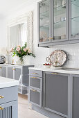 Pale grey kitchen counter, wall units with glass doors and vase of roses on sideboard